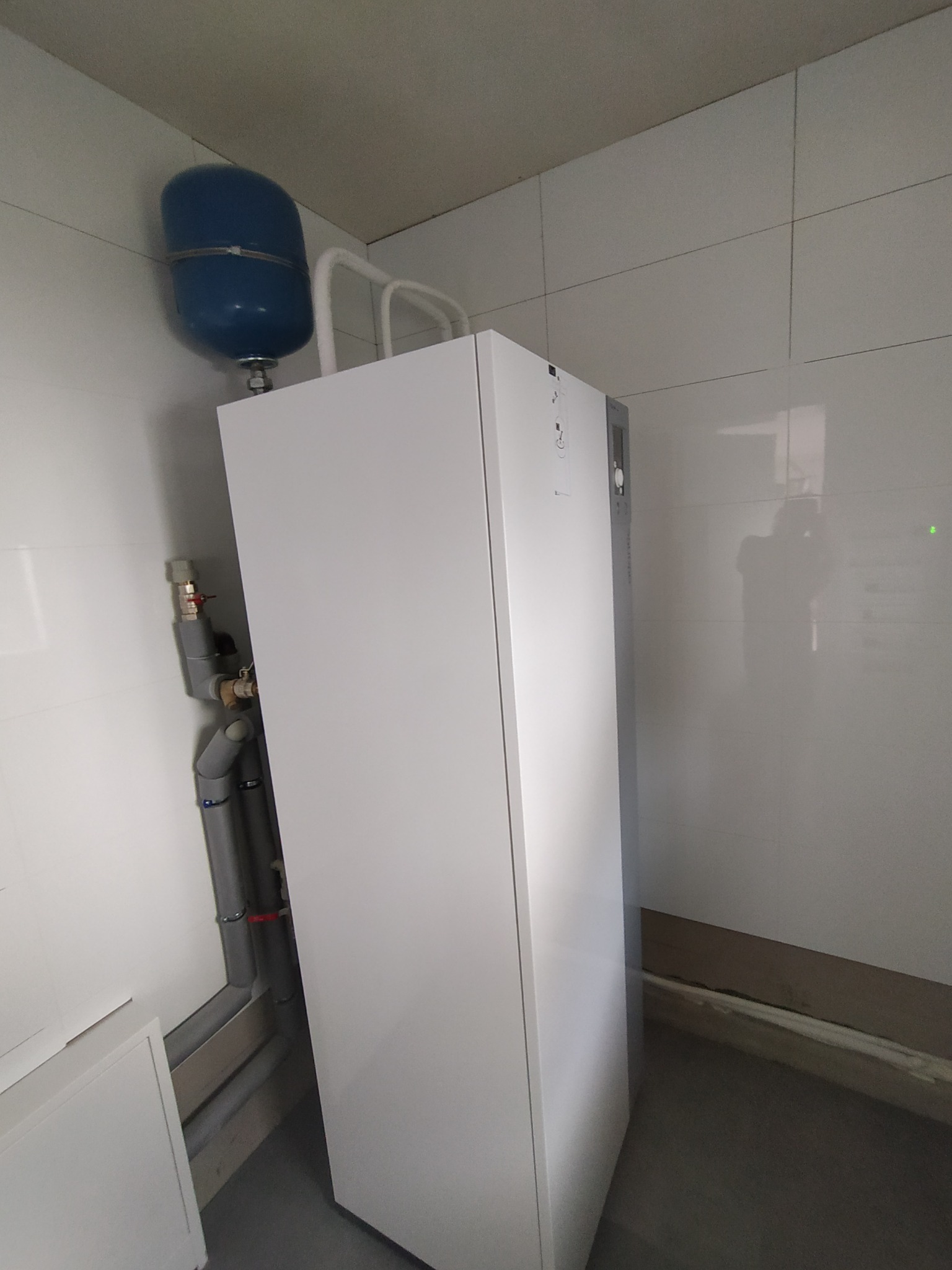 Nowoczesny, biały piec gazowy z niebieskim zbiornikiem wyrównawczym, zamontowany na tle białych płytek ceramicznych, widoczne rury instalacji.