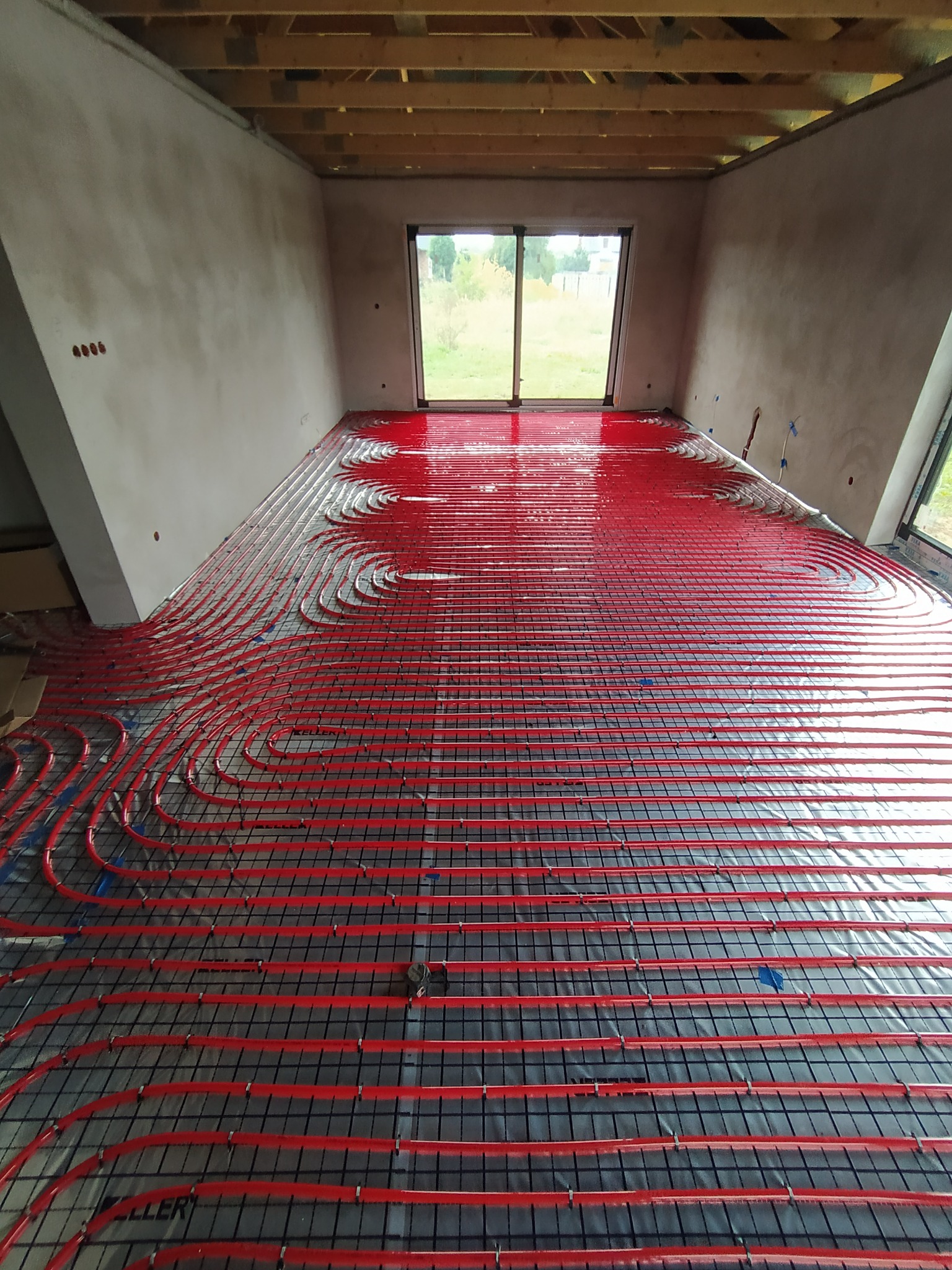 Czerwone rury ogrzewania podłogowego ułożone na czarnej siatce w surowym pomieszczeniu z widokiem na ogród przez okno.