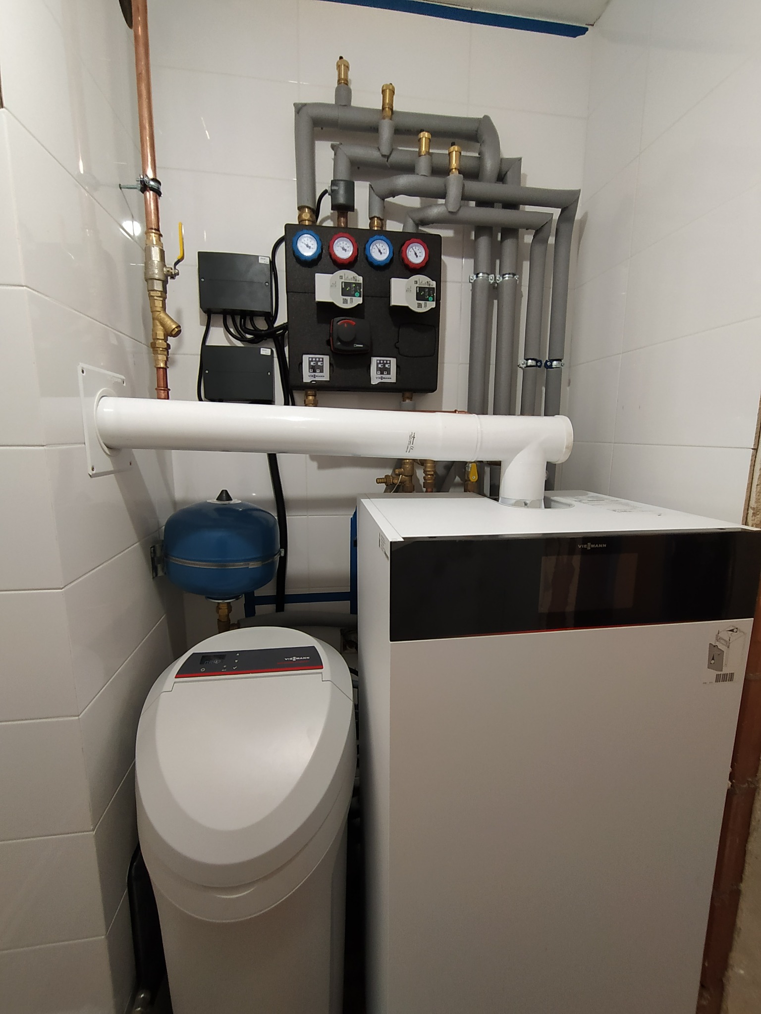 Kompaktowa kotłownia: biały piec Viessmann, szary system rur z izolacją, czarny panel kontrolny z manometrami, niebieski zbiornik wyrównawczy, oraz zmiękczacz wody w pomieszczeniu wyłożonym białymi...