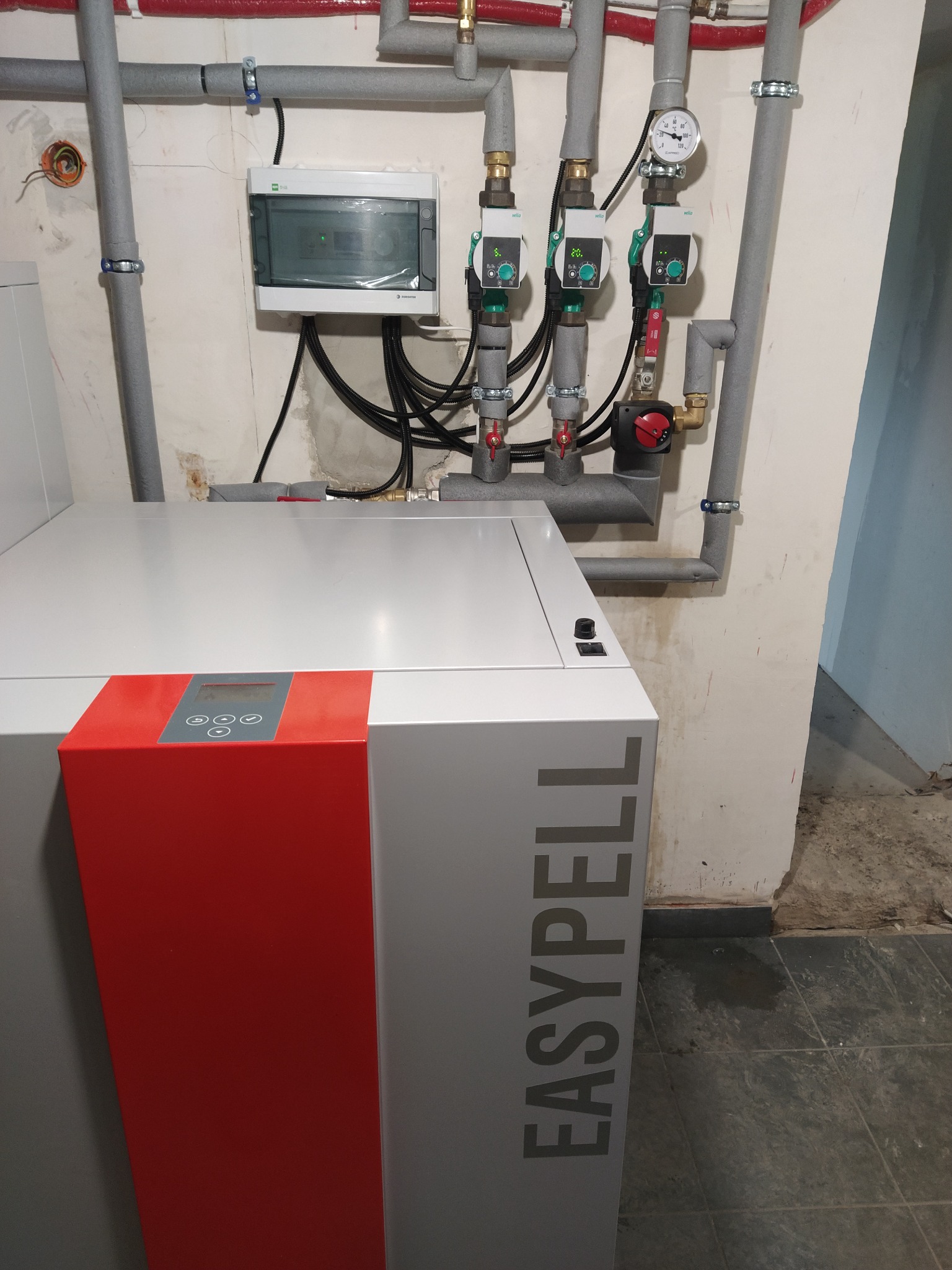 Nowoczesny piec na pellet EasyPell z widocznymi rurami instalacji grzewczej, pompami obiegowymi Wilo i manometrem w kotłowni o surowym wykończeniu.