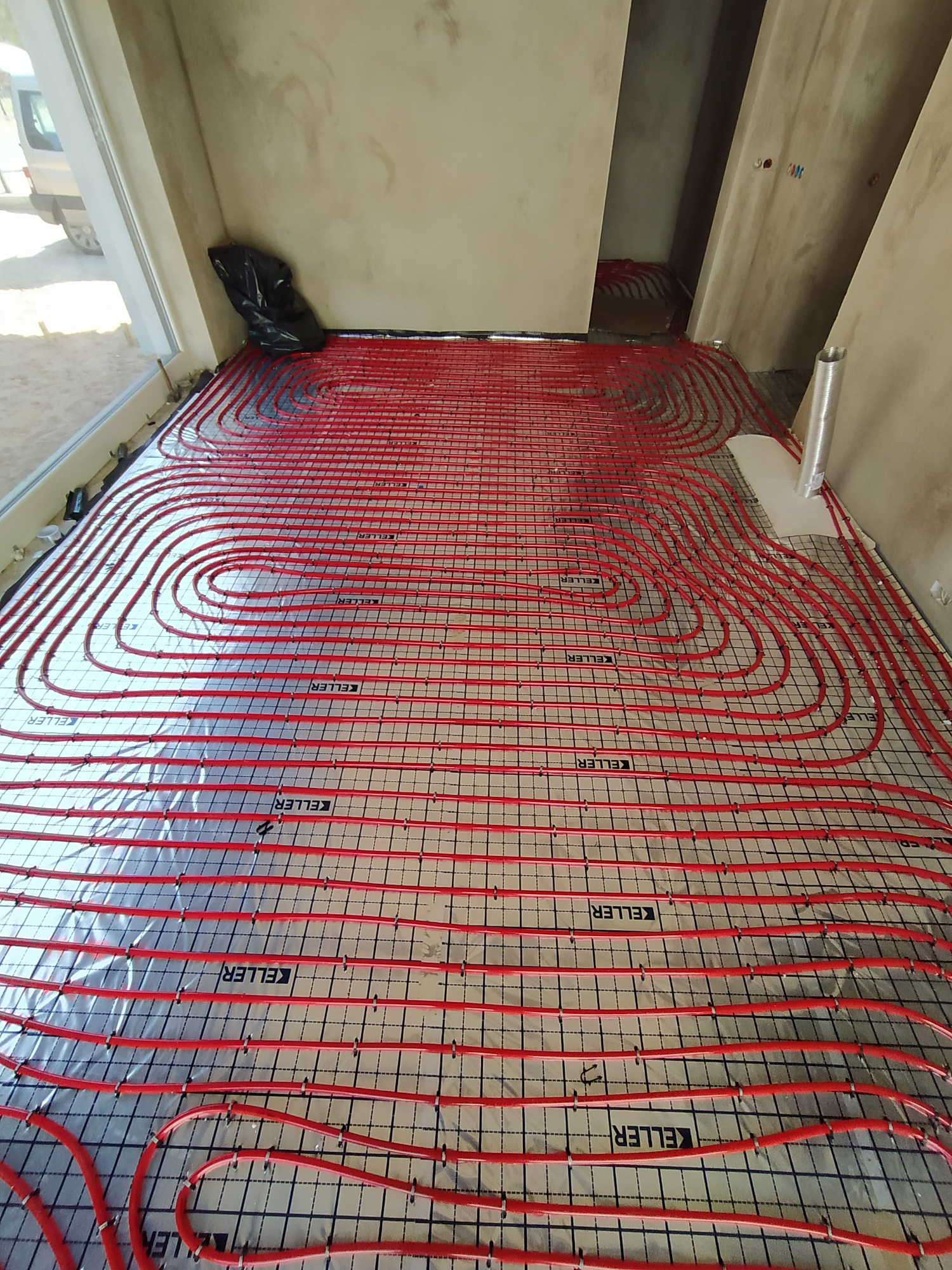 Rozłożone, czerwone rury ogrzewania podłogowego marki Keller na srebrnej folii izolacyjnej z widocznymi zaczepami, przygotowane do zalania wylewką, widok na surowy stan ścian.