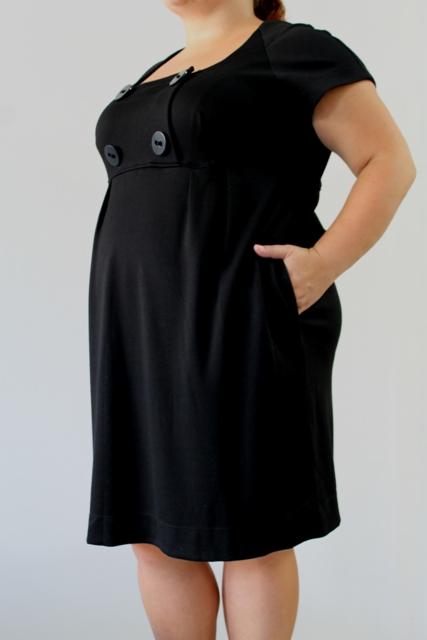Czarna, używana sukienka z krótkim rękawem i ozdobnymi guzikami przy dekolcie, kobieta trzyma rękę w kieszeni.