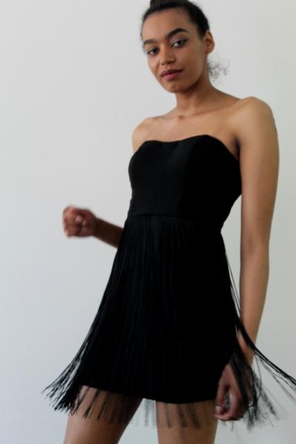 Kobieta w czarnej, bezramiączkowej sukience z frędzlami na białym tle, delikatny ruch w ujęciu.
