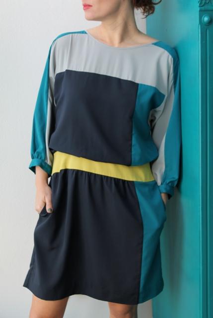 Kobieta w geometrycznej sukience z długim rękawem, o blokach kolorystycznych: szarym, granatowym, turkusowym i żółtym, oparta o turkusowe drzwi.