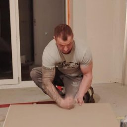 Bud-mat - Mężczyzna w trakcie docinania płyty gipsowo-kartonowej w pomieszczeniu z dużym oknem. Widoczne narzędzia i fragment ściany w trakcie prac wykończeniowych.