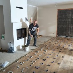 Bud-mat - Układanie drewnopodobnych płytek podłogowych w salonie z kominkiem. Glazurnik uśmiechnięty, w tle zabezpieczone folią drzwi. Widoczne narzędzia i system poziomowania.