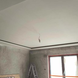 Bud-mat - Wykończony sufit podwieszany z oświetleniem LED w nowo wykończonym pokoju. Widoczne ściany z efektem betonu i okno zabezpieczone folią. Ustawiona drabina.