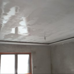 Bud-mat - Wykończony sufit podwieszany z gładzią, widoczne okno i drabina malarska w surowym wnętrzu. Oświetlenie odbija się od gładkiej powierzchni sufitu.