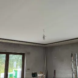 Gładki, biały sufit z dekoracyjną listwą w wykończonym wnętrzu. Ściany przygotowane do malowania. Widoczne okna i drabina malarska.