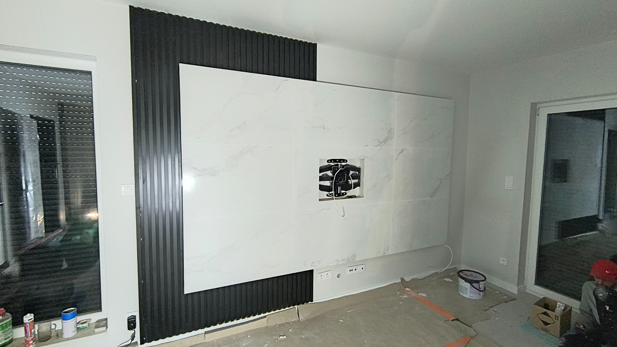 Montaż płytek imitujących marmur i dekoracyjnych lameli na ścianie w salonie. Widoczny uchwyt do telewizora. Prace wykończeniowe w trakcie realizacji.