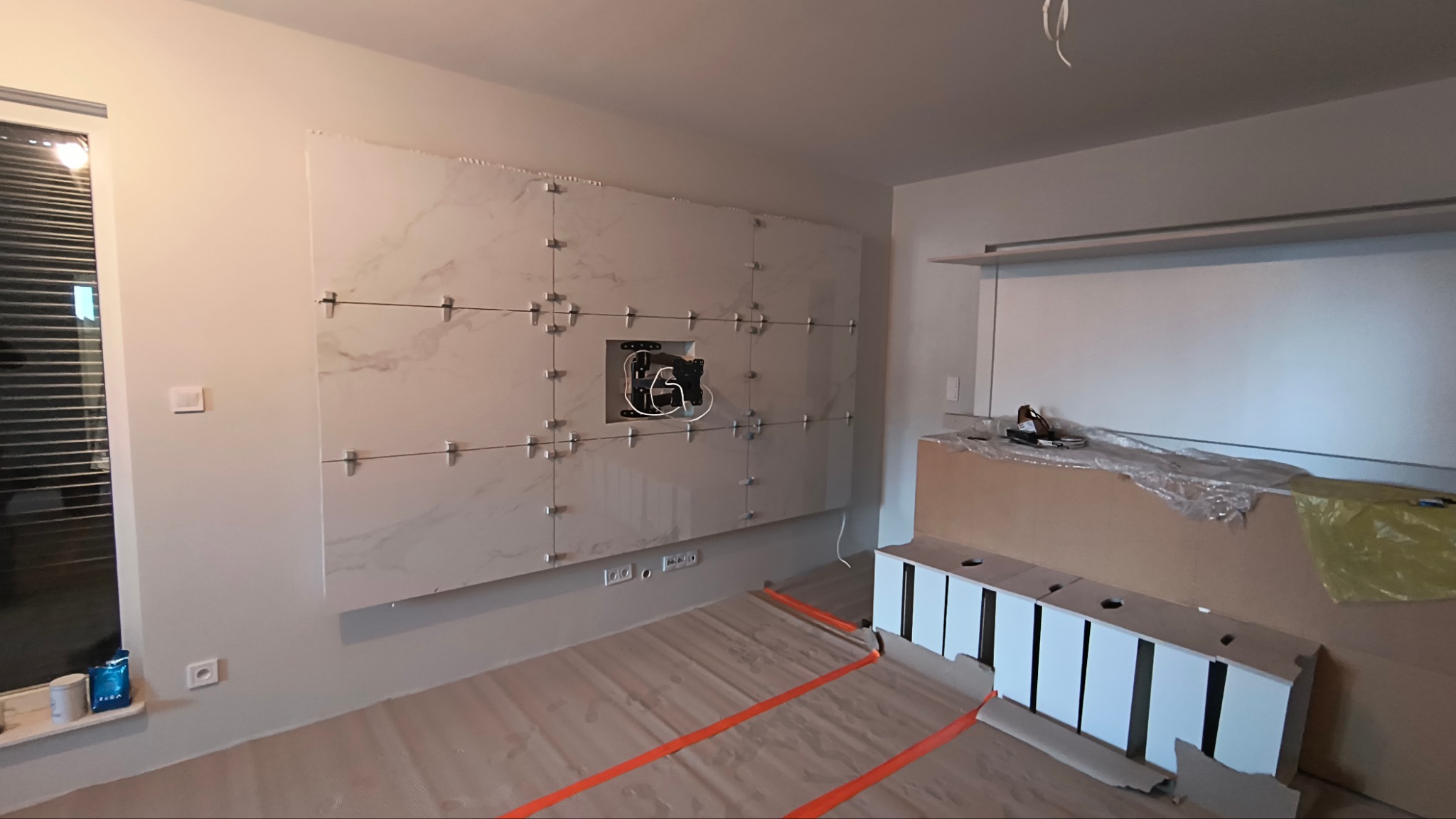 Ściana z jasnych, marmurkowych płytek w trakcie montażu, z widocznymi krzyżykami dystansowymi i uchwytem na telewizor, w nowoczesnym wnętrzu.