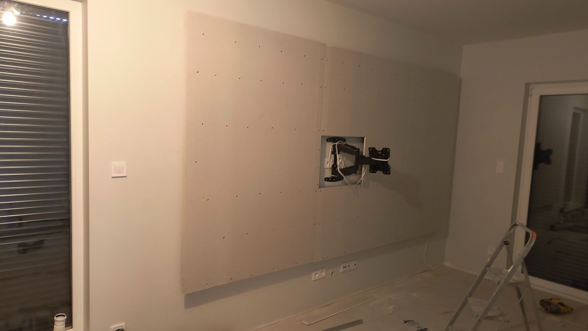 Ściana z płyt gipsowo-kartonowych w trakcie montażu, widoczny uchwyt na telewizor i ślady szpachlowania. W tle okno i drabina.