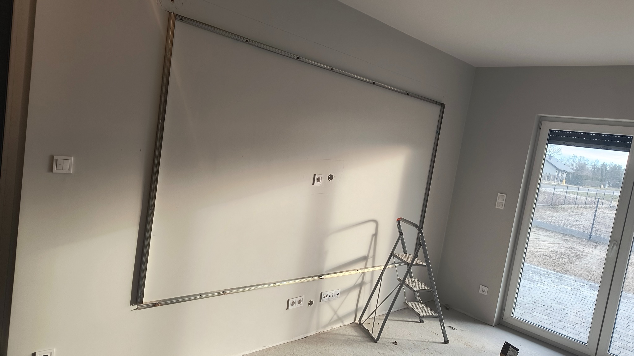 Montaż stelaża pod płytę G-K na ścianie z gładzią, widoczne puszki elektryczne i drabina. W tle okno z widokiem na zewnątrz i fragment posadzki.