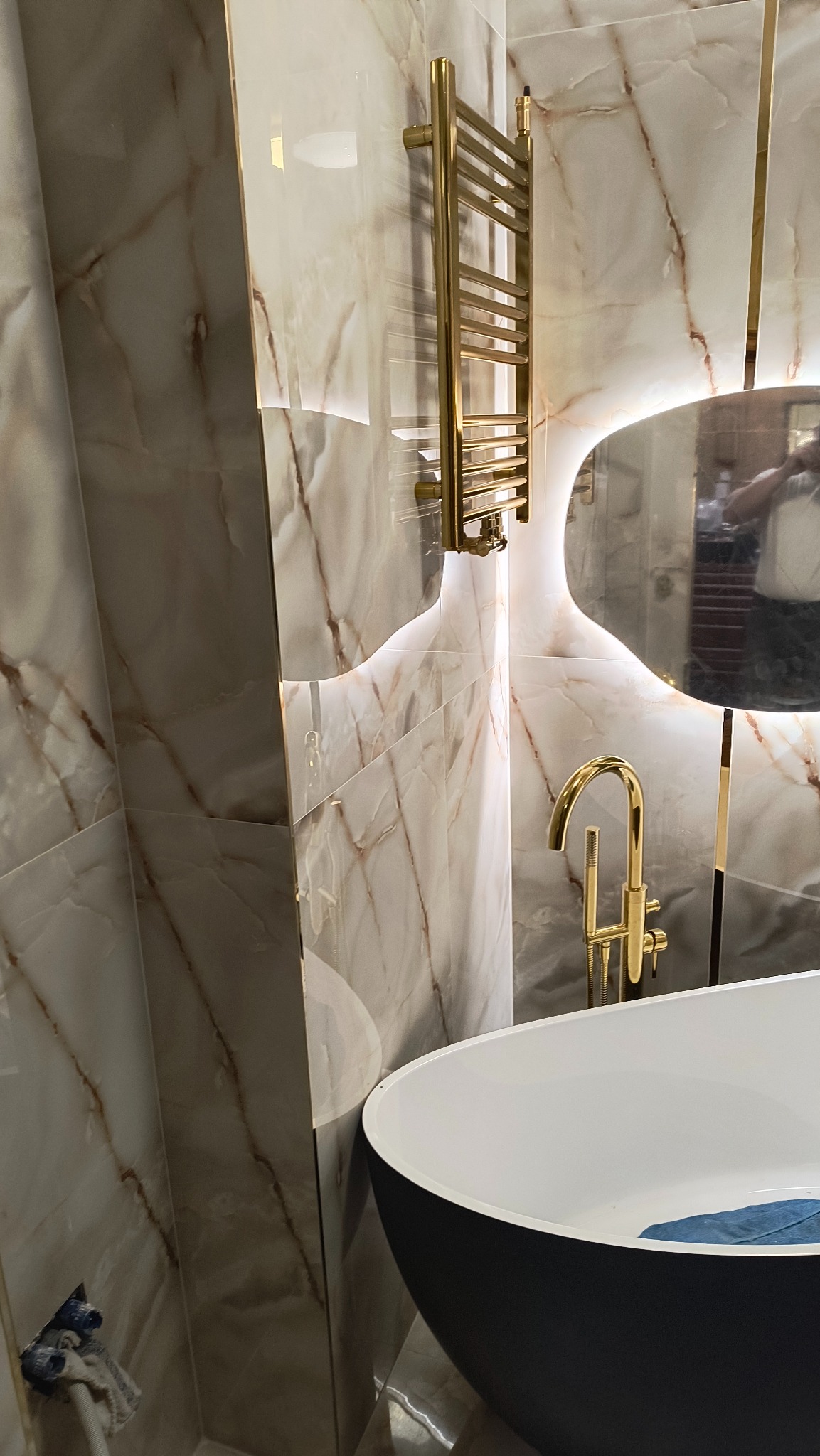 Elegancka łazienka z wanną wolnostojącą, złotą armaturą i marmurowymi płytkami. Oświetlenie LED za lustrem dodaje luksusowego charakteru.