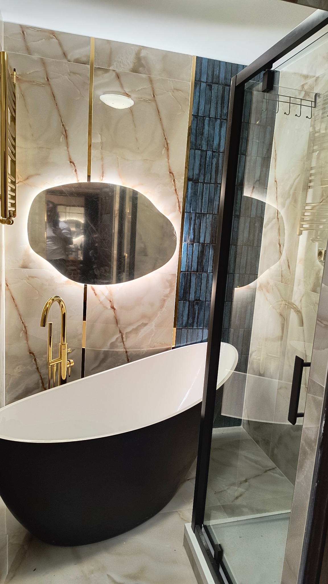 Elegancka łazienka z czarną wanną wolnostojącą, złotą armaturą i podświetlanym lustrem o nieregularnym kształcie na tle marmurowych i niebieskich płytek.