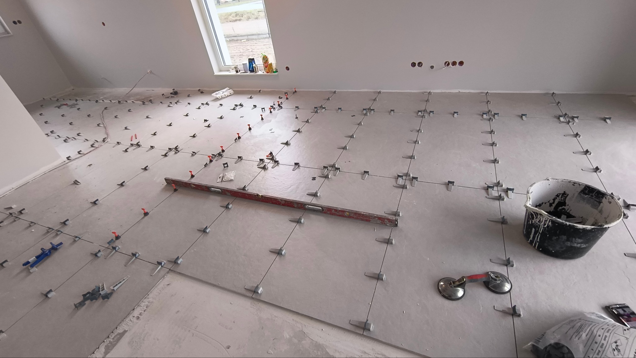 Układanie jasnych płytek podłogowych z systemem poziomowania, widoczny poziomica, przyssawka do płytek i wiadro z zaprawą w jasnym pomieszczeniu.