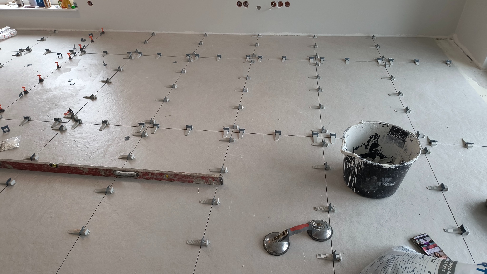 Jasne płytki podłogowe w trakcie układania z użyciem systemu poziomowania, widoczna poziomica, przyssawka do płytek i wiadro po kleju. Delikatne oświetlenie.