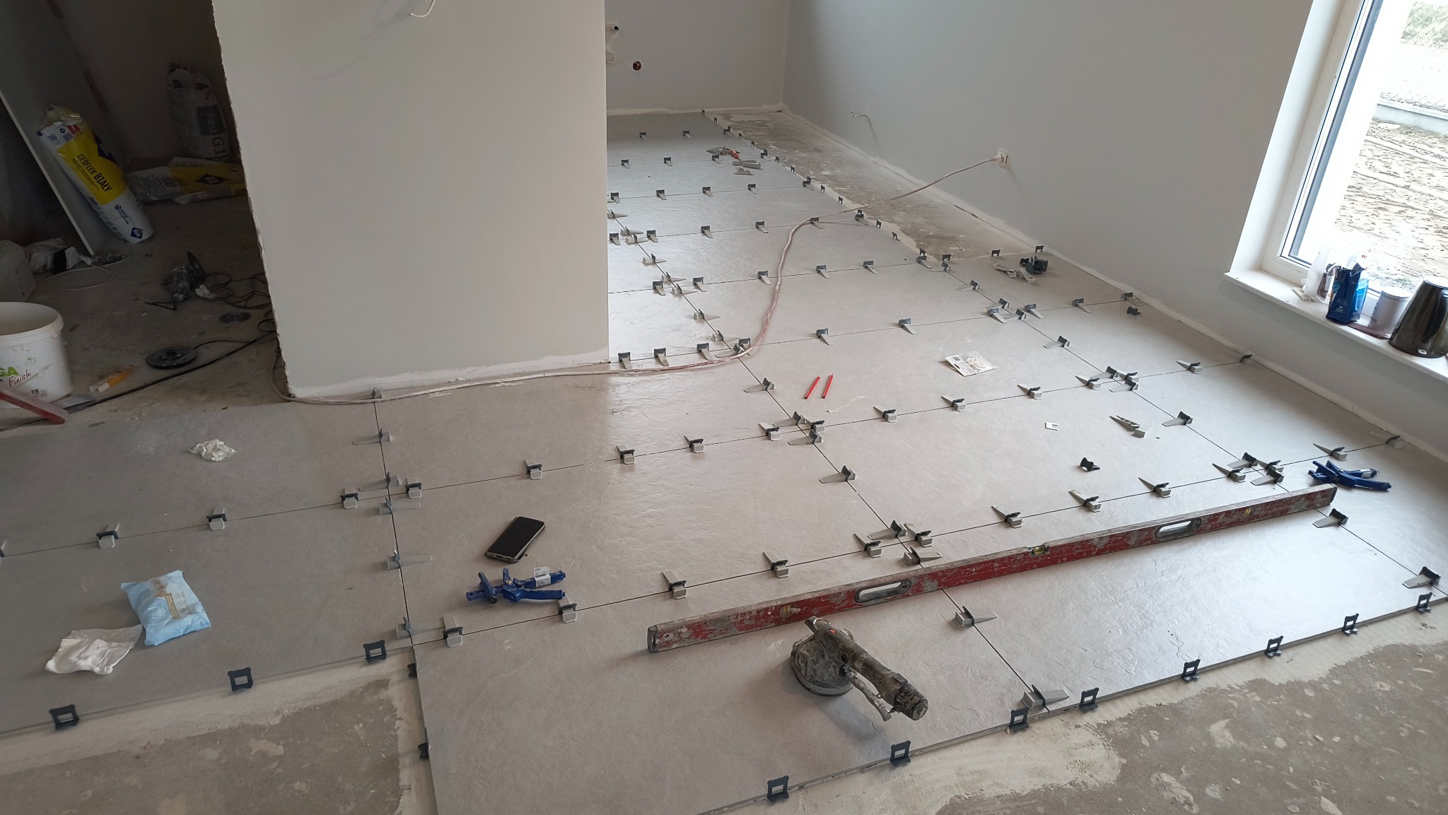 Układanie jasnych płytek podłogowych z użyciem krzyżyków dystansowych. Poziomica, przyssawka do płytek i telefon na podłodze. Widok na okno i sąsiednie pomieszczenie.