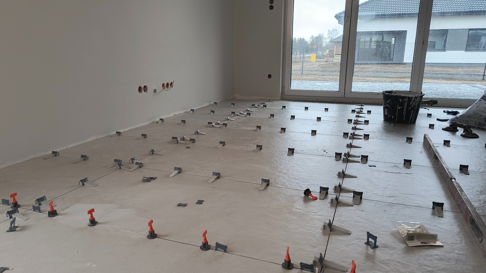 Układanie płytek podłogowych z użyciem systemu poziomowania w Wygodzie. Widoczne krzyżyki dystansowe i kliny, przygotowane podłoże i narzędzia glazurnicze.