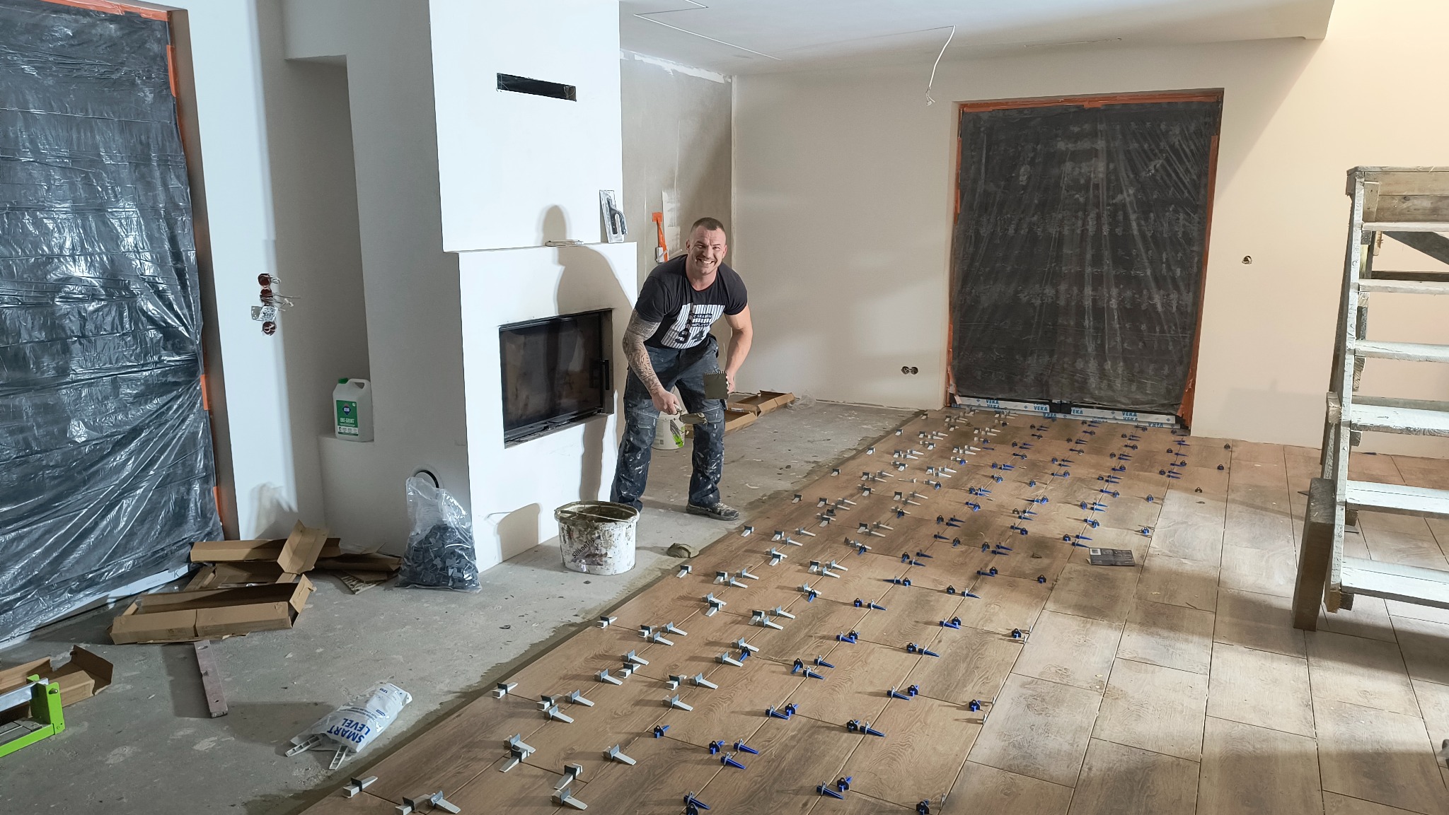 Układanie drewnopodobnych płytek podłogowych w salonie z kominkiem. Glazurnik uśmiechnięty, w tle zabezpieczone folią drzwi. Widoczne narzędzia i system poziomowania.