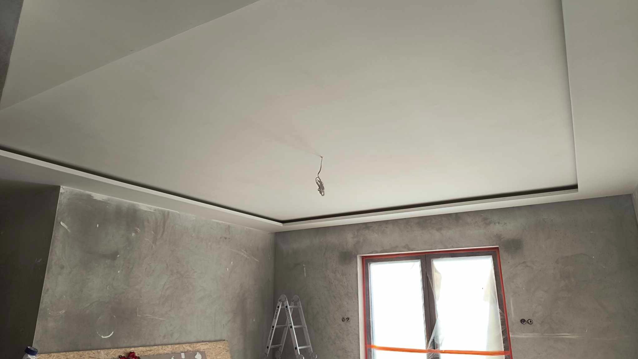 Wykończony sufit podwieszany z oświetleniem LED w nowo wykończonym pokoju. Widoczne ściany z efektem betonu i okno zabezpieczone folią. Ustawiona drabina.