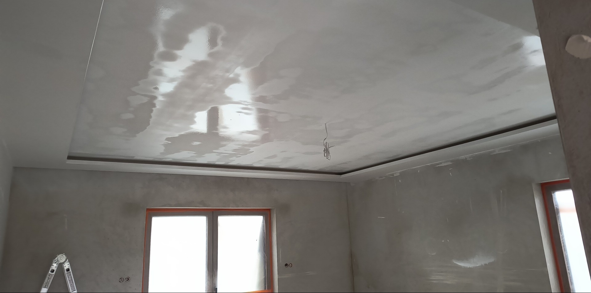 Wykończony sufit podwieszany z gładzią, widoczne okno i drabina malarska w surowym wnętrzu. Oświetlenie odbija się od gładkiej powierzchni sufitu.
