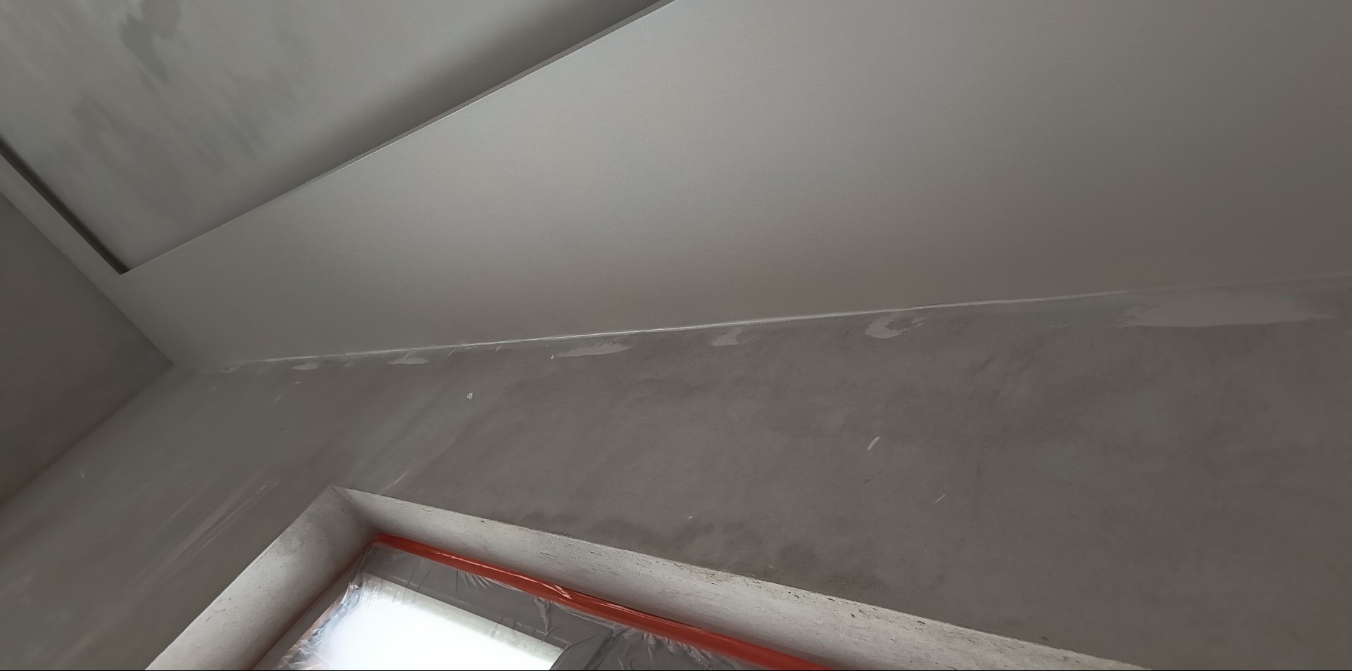 Szpachlowana ściana i sufit podwieszany z płyty gipsowej, zabezpieczone okno folią. Przygotowanie do malowania w trakcie remontu.