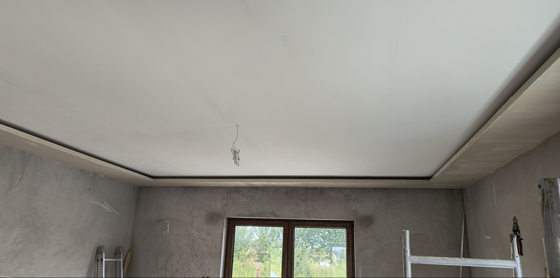 Biały sufit podwieszany z oświetleniem w surowym wnętrzu. Widoczne ślady szpachlowania na ścianach i wystające kable. Fragment drabiny malarskiej.