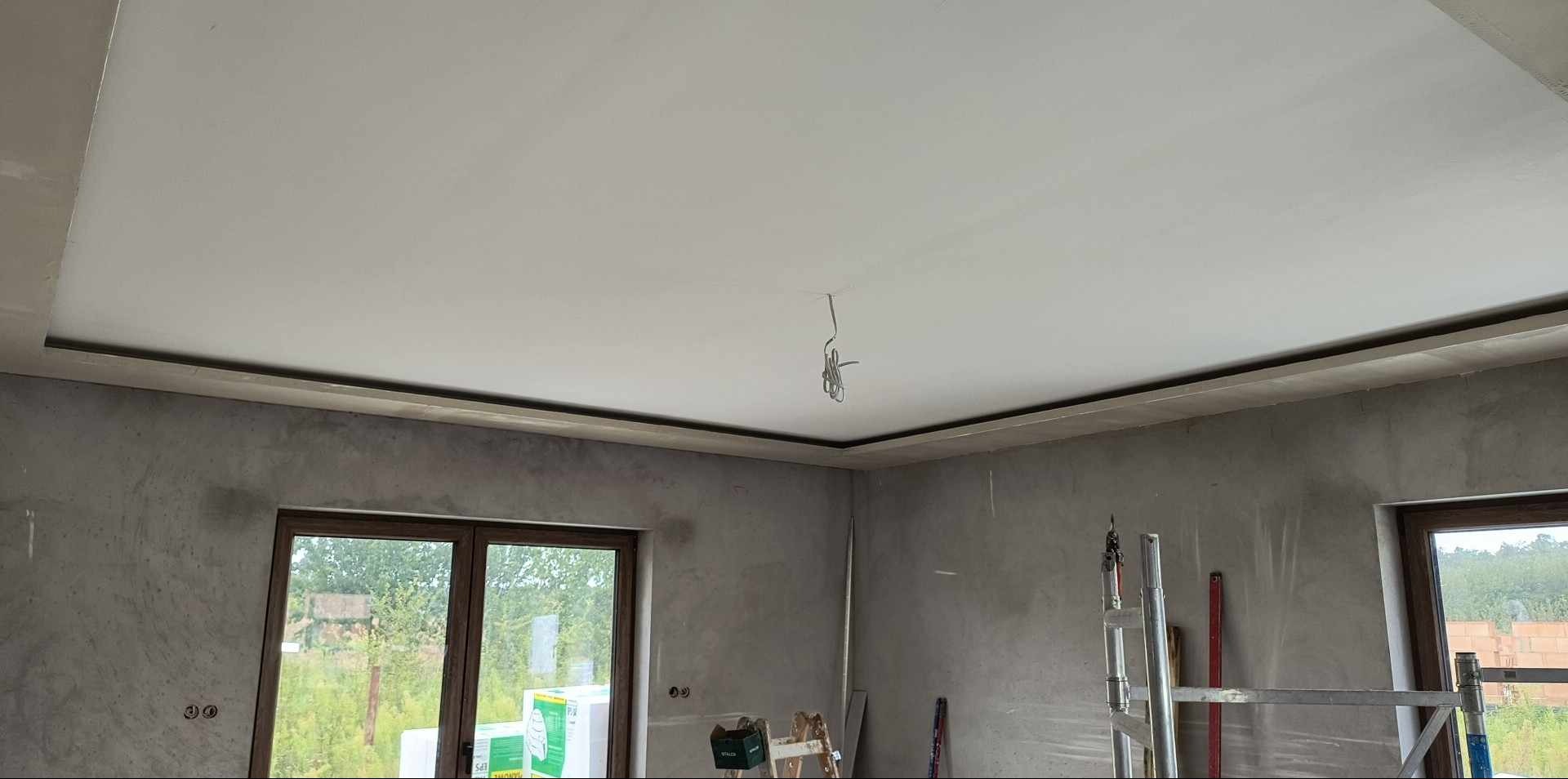 Gładki, biały sufit z dekoracyjną listwą w wykończonym wnętrzu. Ściany przygotowane do malowania. Widoczne okna i drabina malarska.