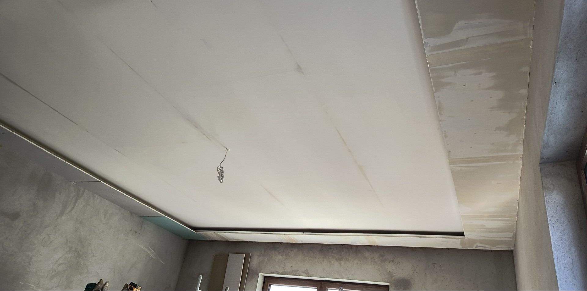 Sufit podwieszany z płyt gipsowo-kartonowych w trakcie szpachlowania, widoczne łączenia płyt i wystające przewody elektryczne. Surowe ściany w tle.