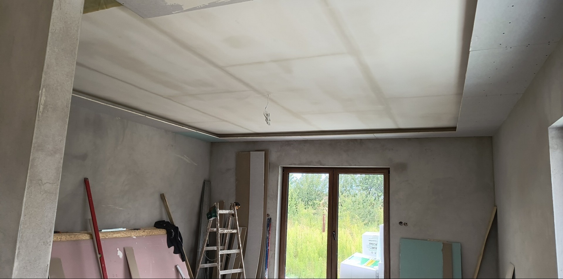 Sufit podwieszany z płyt kartonowo-gipsowych w trakcie montażu, widoczne szpachlowane łączenia, surowe ściany i elementy konstrukcyjne. W tle okno z widokiem na zieleń.