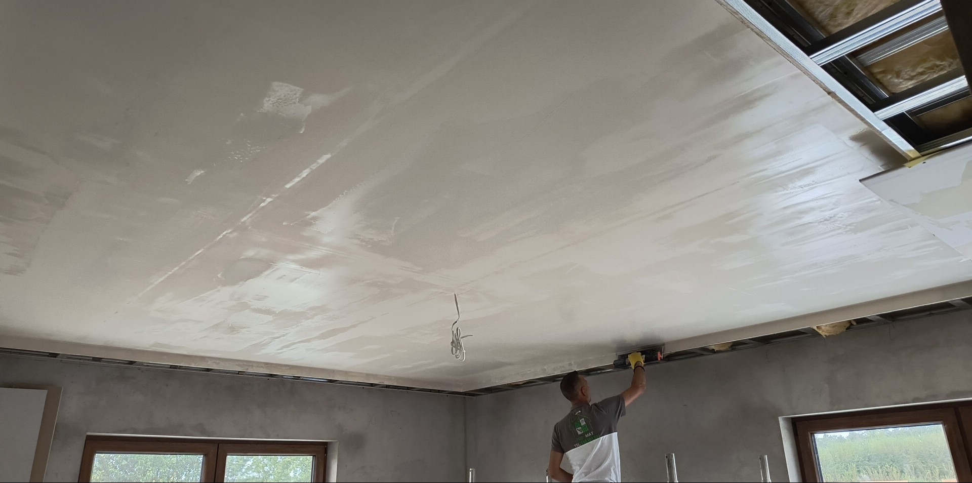 Praca na wysokości: szpachlowanie sufitu z płyty gipsowej w pomieszczeniu z oknami. Widoczne metalowe profile i wystające kable. Pracownik w trakcie szlifowania gładzi.