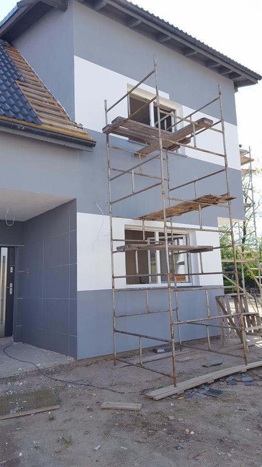 Dom w trakcie malowania elewacji w odcieniach szarości i bieli, widoczne rusztowania i częściowo zdjęte dachówki.