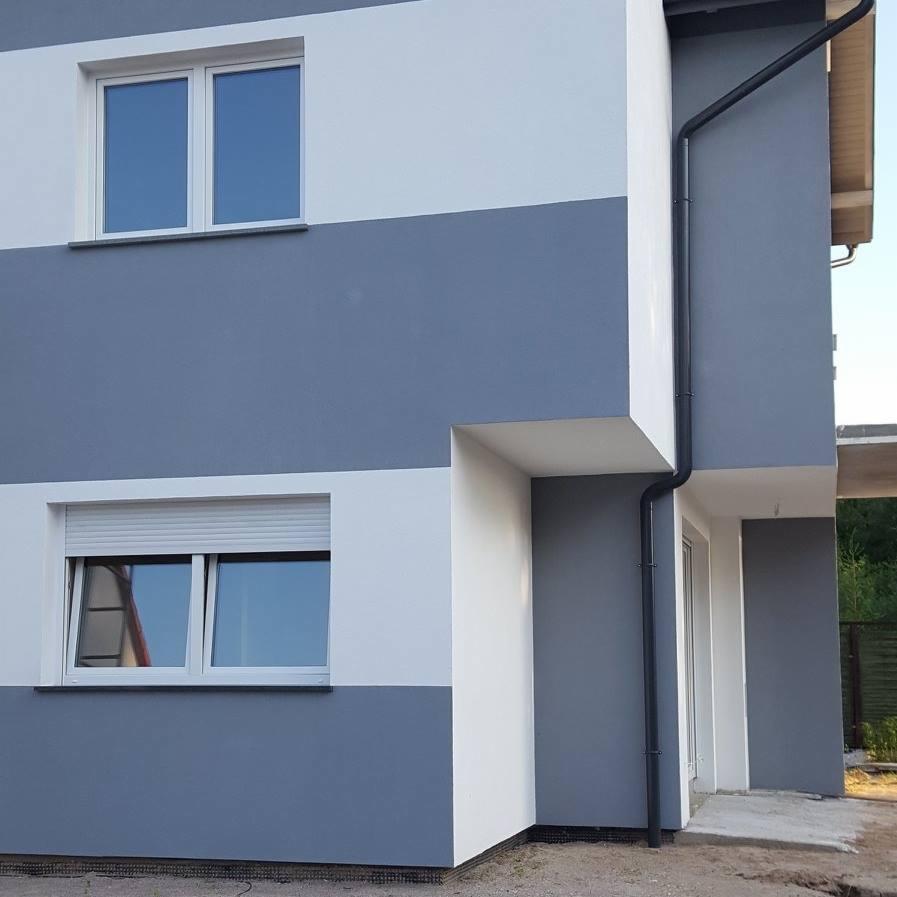 Dwukolorowa elewacja budynku z oknami, szara dolna część i biała górna, z widoczną rynną. Okno na dole z roletą częściowo opuszczoną.