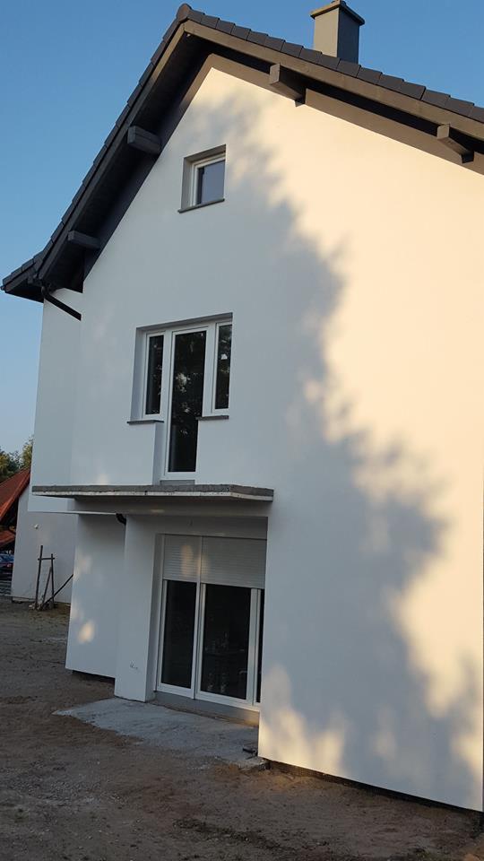 Świeżo otynkowana, biała elewacja dwukondygnacyjnego domu z szarym dachem i kominem, widoczne okna z białymi ramami, rolety zewnętrzne na parterze, oraz cień rzucany przez drzewo na ścianę.