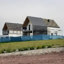 Dwa domy w trakcie budowy, jeden z szarą blachą na dachu, drugi z widoczną konstrukcją dachu, oba zabezpieczone niebieską siatką na tle zielonego wzgórza i pochmurnego nieba.