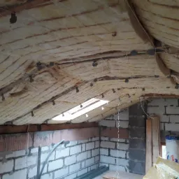 Poddasze w trakcie prac izolacyjnych w Chełmży, widoczna wełna mineralna zamocowana do krokwi, fragment ściany z pustaków oraz okno dachowe.