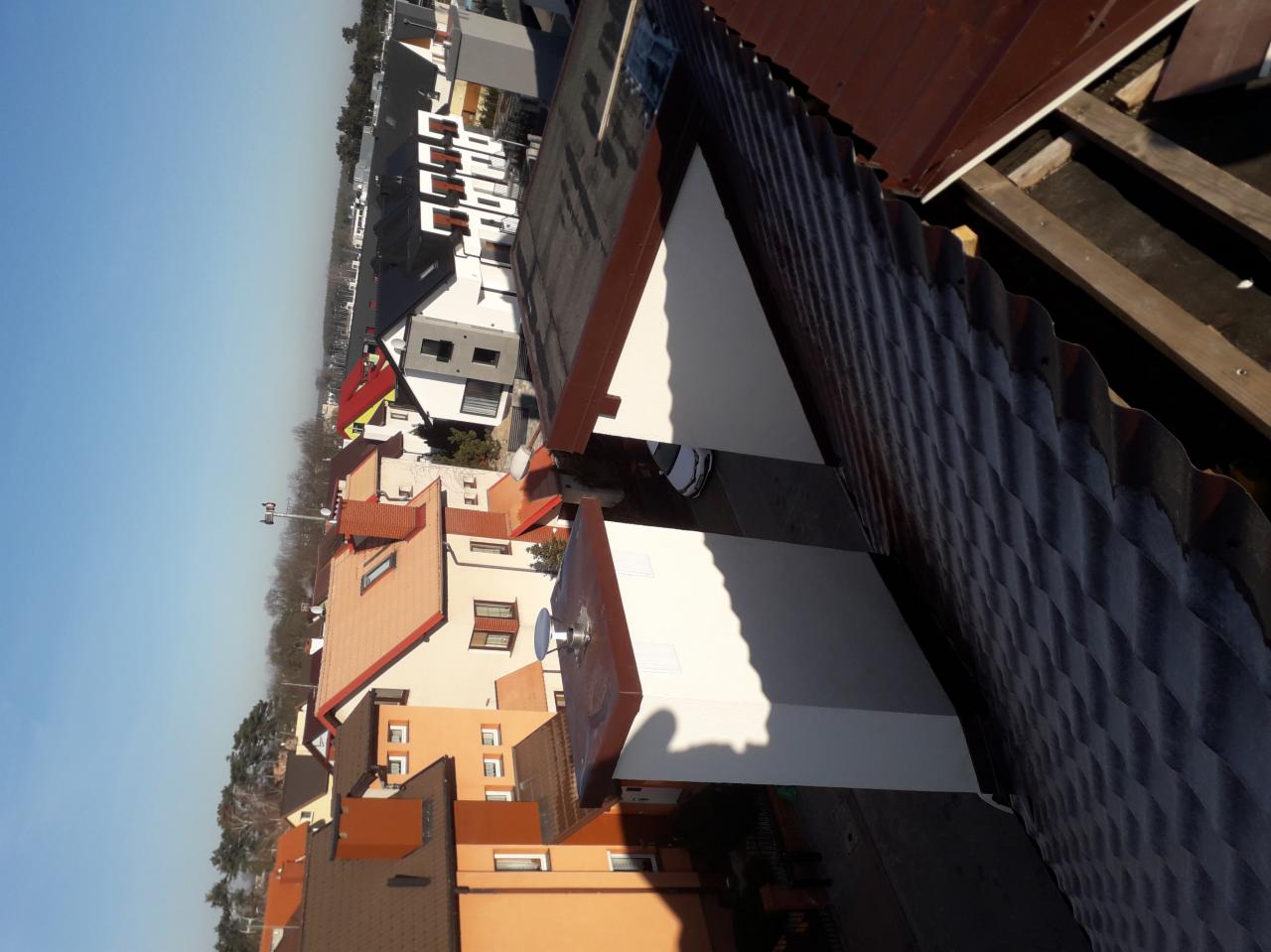 Widok z góry na fragment dachu z blachy falistej w trakcie montażu, z widoczną konstrukcją więźby i fragmentem białej ściany, w tle zabudowa miejska z różnorodnymi kolorami dachów.