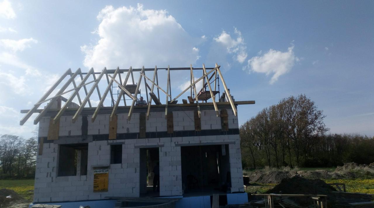 Budowa domu jednorodzinnego w stanie surowym otwartym, widoczna murowana konstrukcja ścian z betonu komórkowego oraz drewniana konstrukcja dachu bez pokrycia, na tle błękitnego nieba z chmurami.