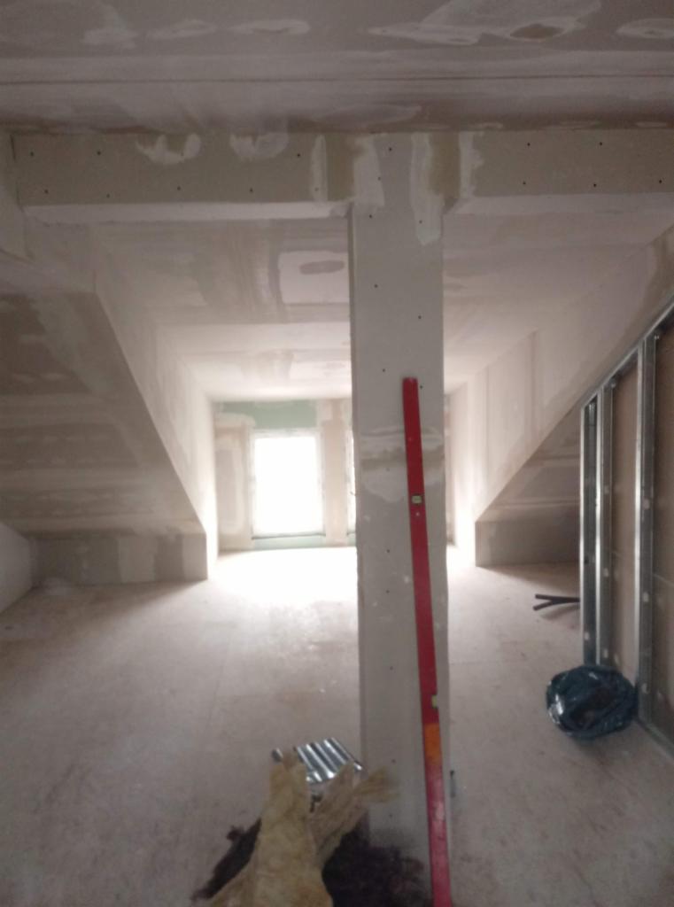 Poddasze w trakcie wykańczania płytami gipsowo-kartonowymi, widoczny pionowy profil z czerwoną poziomicą, fragment izolacji i metalowe profile ścienne.