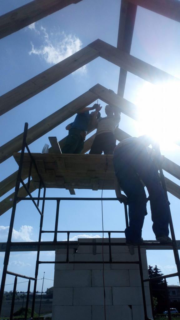 Trzech dekarzy montuje więźbę dachową na rusztowaniu, widok z dołu na tle błękitnego nieba z pojedynczymi chmurami.