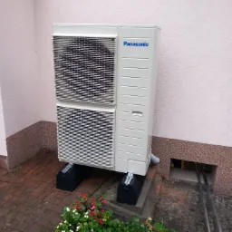 Biała, dwuczęściowa jednostka zewnętrzna z niebieskim logo Panasonic, umieszczona na czarnych, podłużnych podstawach na tle różowej ściany budynku i ceglanej kostki brukowej, z widocznymi rurami...