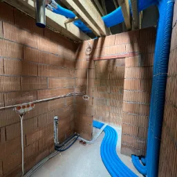 Wnętrze w trakcie budowy z widocznymi niebieskimi rurami wentylacyjnymi pod sufitem i wzdłuż ściany, instalacja elektryczna w peszlach na ścianach z cegły dziurawki.