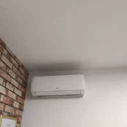 Biała klimatyzacja marki AUX zamontowana na białej ścianie, obok fragmentu ściany z cegły w odcieniach czerwieni i brązu. Widoczny fragment dyplomu w złotej ramce.