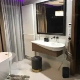 Nowoczesna łazienka z owalną umywalką nablatową, lustrem w czarnej ramie, białymi płytkami podłogowymi z delikatnym wzorem, szarym pufem i akcentami fioletowego oświetlenia za zasłoną.