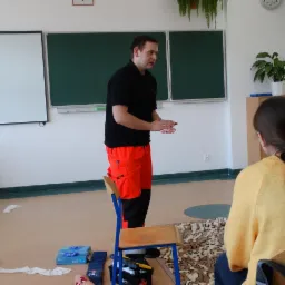 Instruktor w czarnej koszulce i pomarańczowych spodniach demonstruje technikę medyczną przed grupą osób siedzących na krzesłach w sali z tablicą i sprzętem do udzielania pierwszej pomocy.