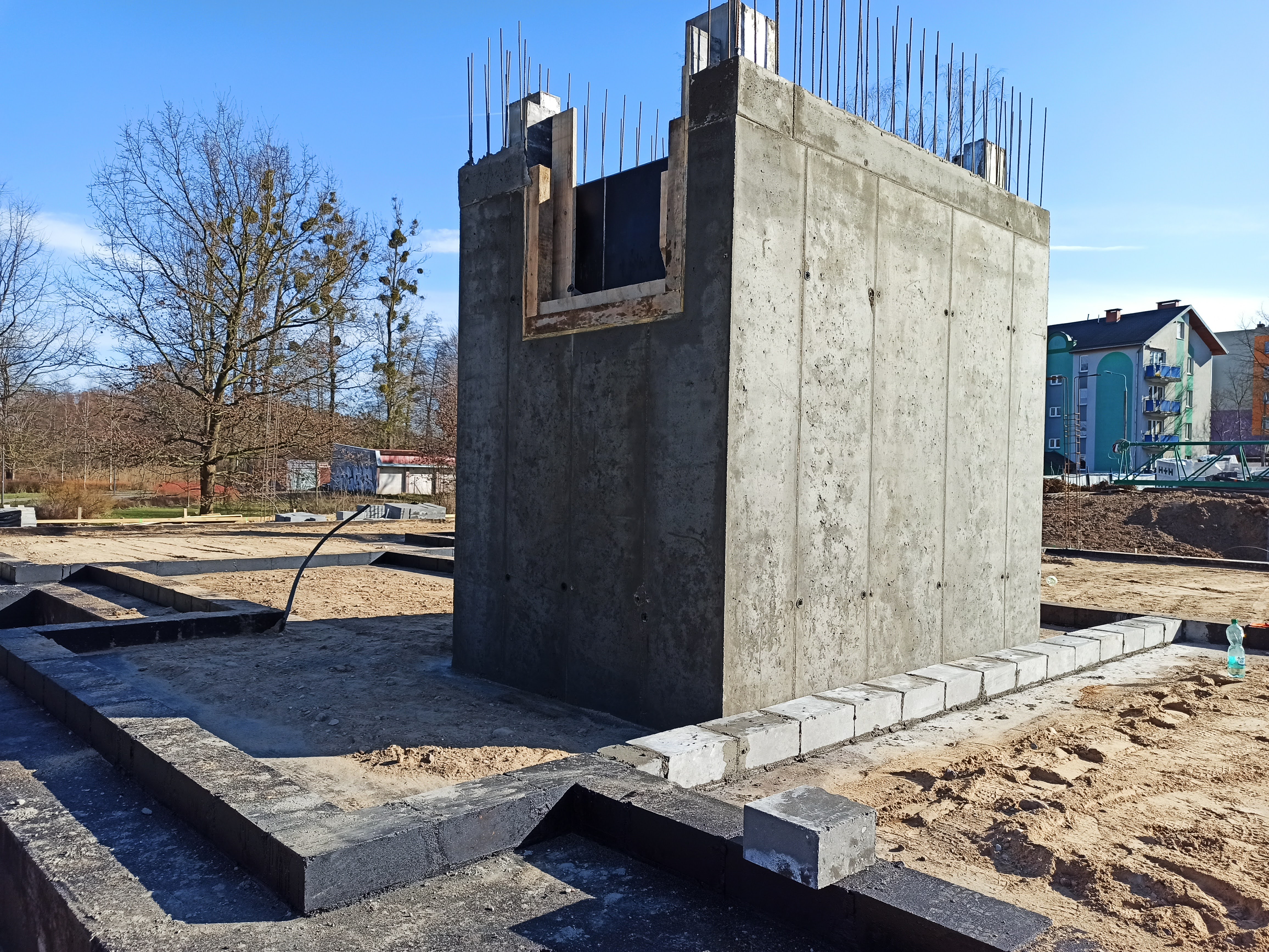 Wznoszenie betonowej konstrukcji z wystającymi prętami zbrojeniowymi na placu budowy, widoczne fundamenty i szalunki okienne, w tle drzewa i budynek mieszkalny.