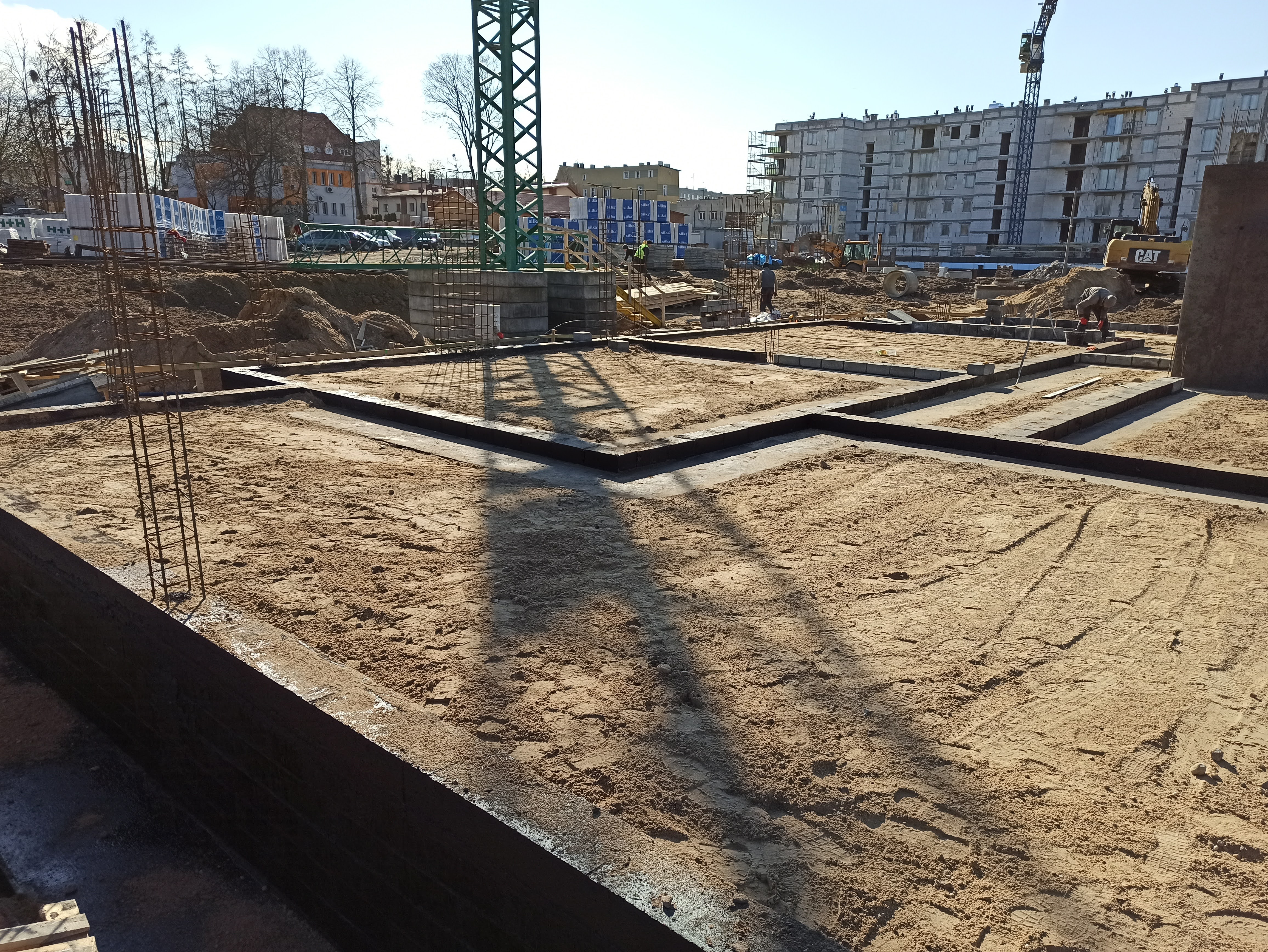 Przygotowanie terenu pod fundamenty budynku wielorodzinnego, widoczne zbrojenie, deskowanie i ubity piasek, w tle dźwig budowlany i budynek w stanie surowym.