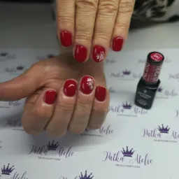Dłonie z czerwonym manicure hybrydowym, zdobione białymi kwiatowymi wzorami na tle firmowego papieru z logo i buteleczką lakieru.