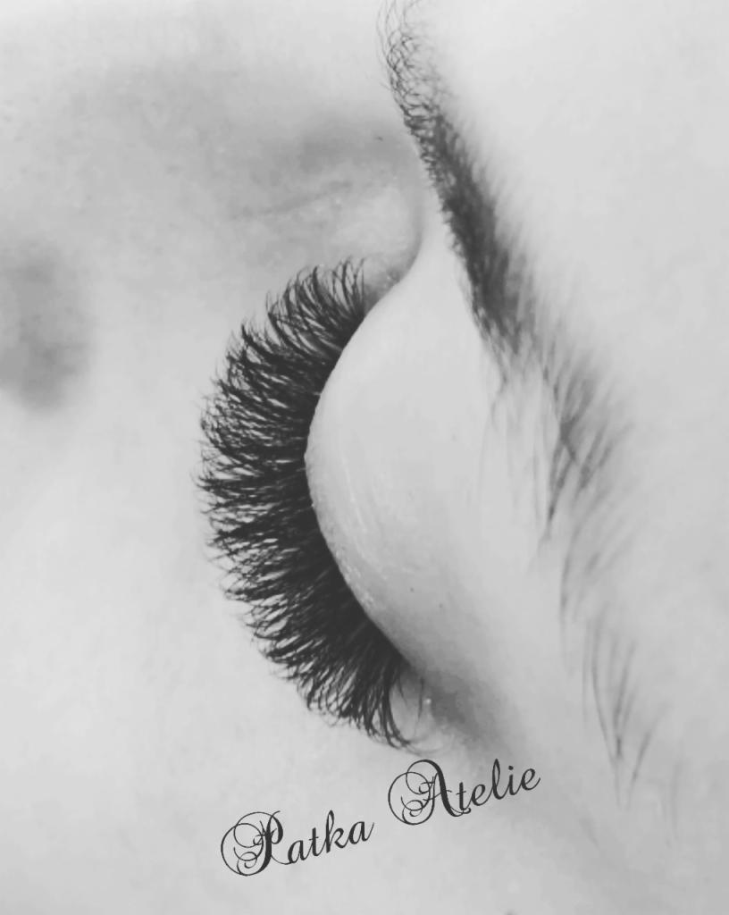 Czarno-białe zbliżenie na oko z gęstymi, przedłużonymi rzęsami, widoczny delikatny makijaż brwi, w dolnym rogu logo 'Patka Atelie'.